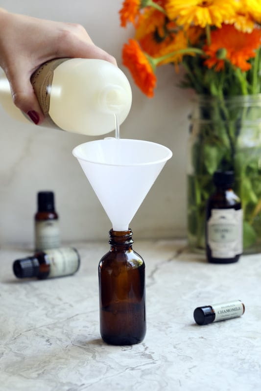 Hand pouring liquid through funnel into amber class jar with bottles of oil in the background