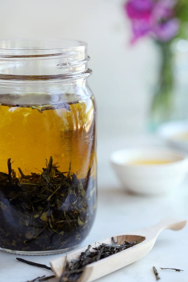 A mason jar filled with green tea leaves and organic olive oil left to steep. A wooden scoop in the foreground is filled with green tea leaves and bowls of oil in the background. 