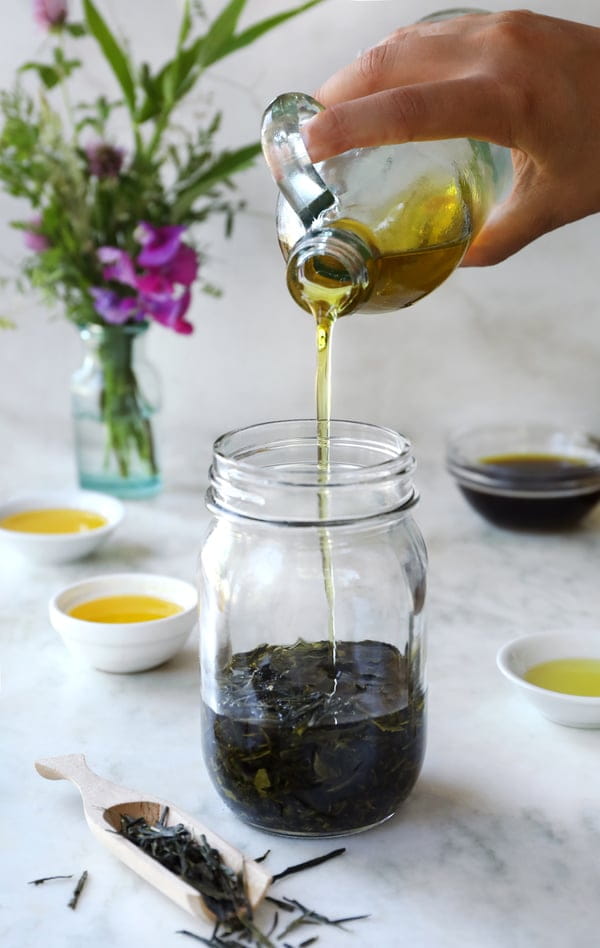 Organic olive oil being poured over dried green tea leaves in a clear mason jar. Bowls of different colored oils are arranged around with a wooden scoop of tea leaves on a white marble counter top. 