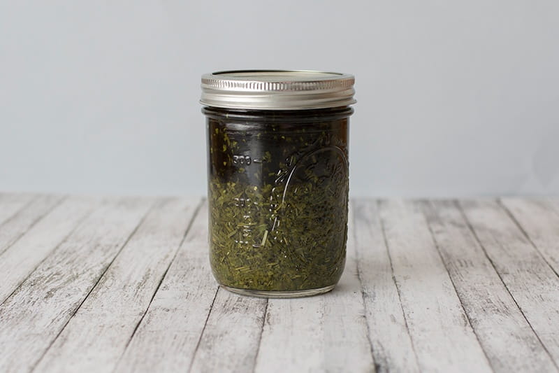 Glass jar full of herbs tincturing on a wooden table with white background.